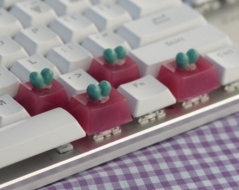 以下が含まれることがあります： 白いキーボードに緑の葉のデザインが施されたピンクのキーキャップが4つあります。キーキャップはF、J、K、Lキーにあります。