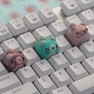 May include: Three small, colorful, animal-shaped keycaps for a computer keyboard. The keycaps are in the shape of a pig, a bear, and a turtle. The pig is pink and purple, the bear is pink, and the turtle is blue and green.