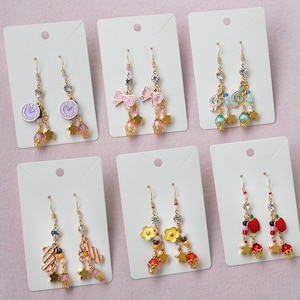 May include: Six pairs of dangle earrings displayed on white cards. The earrings feature a variety of charms, including clocks, bows, musical notes, flowers, and apples. The charms are made from gold-toned metal and are accented with pink, blue, and red beads.