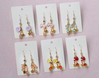 May include: Six pairs of dangle earrings on white card displays. The earrings feature various charms, including clocks, bows, musical notes, flowers, and apples. The charms are made of gold-toned metal and are accented with pink, blue, and red beads.