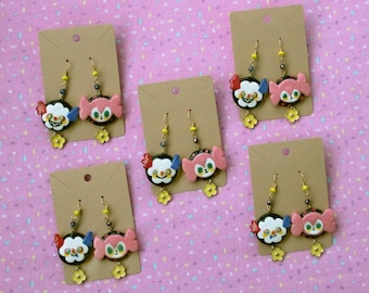 May include: Three pairs of dangle earrings with colorful cartoon faces. Each pair features a black and white face with a red nose and a pink face with a yellow bow. The earrings are on brown card stock.