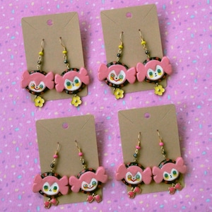 May include: Four pairs of dangle earrings with pink and black candy-like charms. Each charm has a white face with black eyes and a pink mouth. The earrings are displayed on brown card stock.