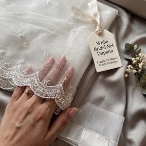Può includere: Un velo da sposa bianco in rete con un delicato bordo ricamato floreale. L'etichetta recita "White Bridal Net Dupatta, Lunghezza: 2,5 metri, Larghezza: 106 cm". Il tessuto trasparente è piegato e legato con un nastro.