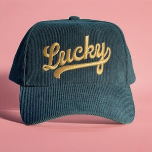 以下が含まれることがあります： 前面に金色の筆記体で「Lucky」の文字が刺繍された、ティール色のコーデュロイベースボールキャップ。キャップにはカーブしたつばと、上部に小さなボタンが付いています。背景は無地のピンク色です。