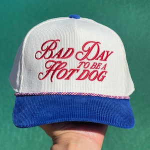 May include: A white corduroy baseball cap with a blue corduroy brim and a red, white, and blue braided rope trim. The front features red embroidered text that reads "BAD DAY TO BE A HOT DOG" in a stylised script and serif font combination.