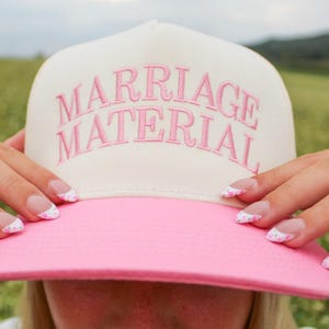 May include: A white and pink trucker hat with the text "MARRIAGE MATERIAL" embroidered in pink on the front.