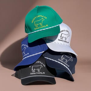 May include: A stack of five baseball caps in green, white, blue, navy, and black. Each cap features a white or yellow embroidered outline of a cow, the text Wood's Cattle Co., and Starke County, IN, with a decorative braided cord on the brim.