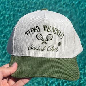 May include: A white and green corduroy baseball cap with the text "Tipsy Tennis Social Club" embroidered on the front. The cap features a green brim and two crossed tennis rackets in the logo.