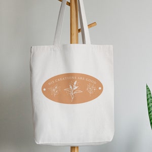 May include: A white canvas tote bag with a brown oval design that says "His Creations Are Good" with a stylized floral design inside the oval.