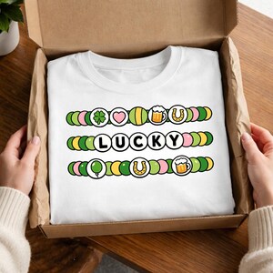 Peut inclure: T-shirt blanc avec le mot "LUCKY" au centre et trois rangées de graphiques colorés au-dessus et en dessous. Les graphiques incluent un trèfle à quatre feuilles, un cœur, une chope de bière, un fer à cheval et un ballon.