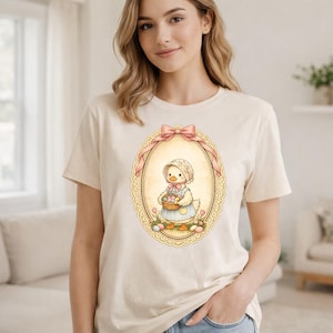 Peut inclure: T-shirt crème avec une illustration encadrée d'un caneton de dessin animé portant un bonnet et un tablier, tenant un panier d'œufs de Pâques. Le cadre a un nœud rose et une bordure en dentelle décorative. Le t-shirt est à col rond et à manches courtes.