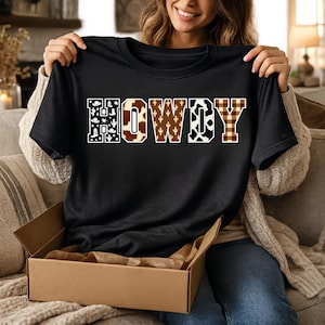 Peut inclure: T-shirt noir avec le mot "HOWDY" dans un design sur le thème du western. Les lettres présentent des motifs tels que des cactus, du cuir de vache et du tartan. Le t-shirt est présenté dans une boîte en carton.