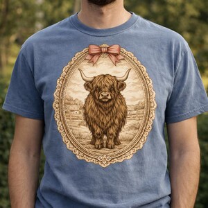 Peut inclure: T-shirt bleu avec une illustration détaillée d'une vache des Highlands dans un cadre ovale. Le cadre est orné d'une bordure décorative et d'un nœud rose. La vache est brune et a de longues cornes.
