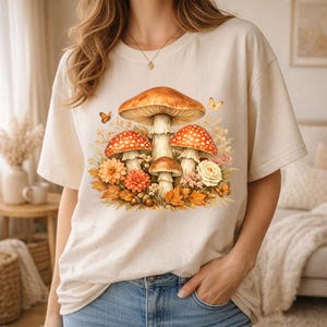 Peut inclure: T-shirt crème avec une illustration détaillée de champignons à chapeaux rouges et blancs tachetés, entourés de fleurs et de feuilles. Le motif comprend des papillons et des couleurs automnales, créant une esthétique inspirée de la nature.