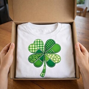 Peut inclure: T-shirt blanc avec un grand motif de trèfle à quatre feuilles vert. Le trèfle est composé de différents motifs verts, dont du vichy, des pois et des imprimés floraux, cousus ensemble. Le t-shirt est présenté dans une boîte en carton ouverte.