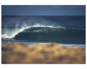 May include: A photograph of a large ocean wave curling over, forming a tube. The wave is a deep teal color, with white foam at the crest. The sky is a dark blue, and the foreground is a blurred sandy color.