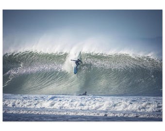 May include: A surfer rides a wave, captured in a dynamic action shot. The surfer, dressed in a dark wetsuit, is balanced on a light blue surfboard, with a massive wave cresting behind them. Another surfer is seen in the water. The ocean is a mix of blues and whites.