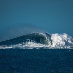 Puede incluir: Una gran ola rompe en el océano, creando una gran cresta espumosa. La ola es de color azul oscuro y el cielo es de color azul claro.