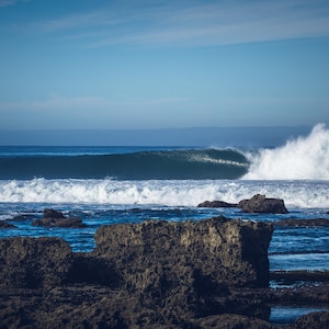 Puede incluir: Una gran ola rompe contra afloramientos rocosos en una playa. La ola se rompe en la distancia, con espuma blanca y agua azul.