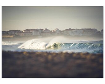 May include: A photograph captures a surfer riding a large, turquoise wave. The wave is breaking, creating a spray of white water. In the background, a coastal town is visible under a hazy sky. The foreground shows a blurred, sandy beach.