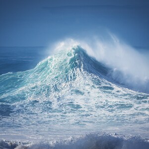 May include: A large, powerful wave crashes in the ocean. The wave is a deep blue color with white foam at the crest. The sky is a clear blue.