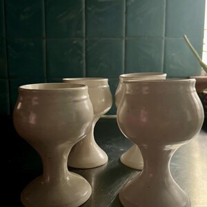 May include: Four off-white ceramic goblets with a bulbous base and a wide rim. The goblets are arranged on a reflective surface, with a teal tile background. The handmade pottery has a rustic, organic aesthetic.