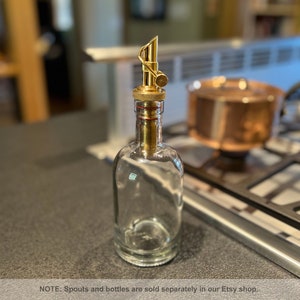 May include: A clear glass bottle with a gold metal spout. The spout has a small knob on the top. The bottle is sitting on a gray countertop.