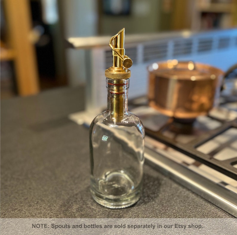 May include: A clear glass bottle with a gold metal spout. The spout has a small knob on the top. The bottle is sitting on a gray countertop.