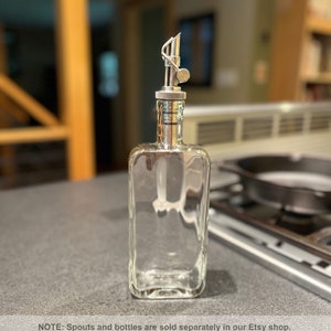 May include: A clear glass bottle with a silver spout and a silver cap. The bottle is empty and sitting on a gray countertop.