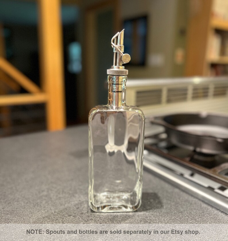 May include: A clear glass bottle with a silver spout and a silver cap. The bottle is empty and sitting on a gray countertop.