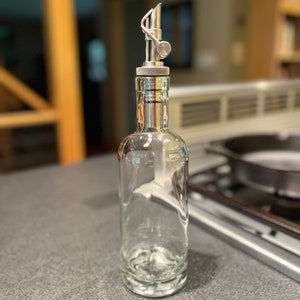 May include: A clear glass bottle with a silver metal spout and a silver metal cap. The bottle is empty and sitting on a gray countertop.
