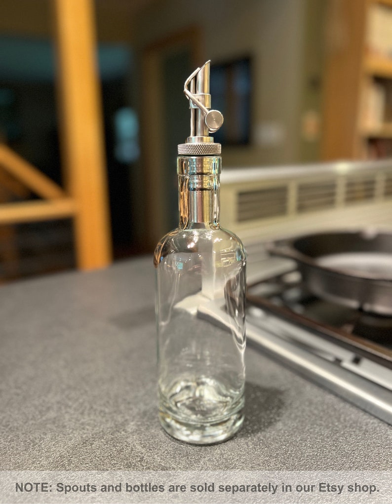 May include: A clear glass bottle with a silver metal spout and a silver metal cap. The bottle is empty and sitting on a gray countertop.