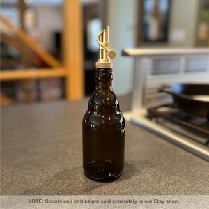May include: A gold metal bottle spout with a curved design and a small handle. The spout is attached to a brown glass bottle.