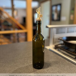 May include: A gold metal bottle spout with a unique design, attached to a dark green glass bottle. The bottle is sitting on a gray countertop.