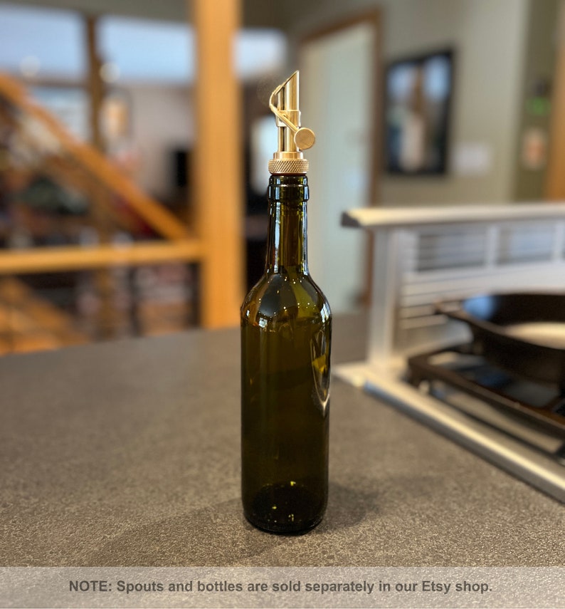 May include: A gold metal bottle spout with a unique design, attached to a dark green glass bottle. The bottle is sitting on a gray countertop.
