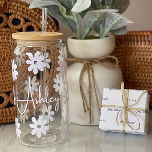 May include: A clear glass water bottle with a bamboo lid and a white straw. The bottle has a white daisy pattern and the name "Ashley" written on it.