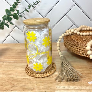 May include: A clear glass jar with a bamboo lid and yellow daisy decals. The jar is sitting on a woven coaster and has the name "Jenny" written on it.