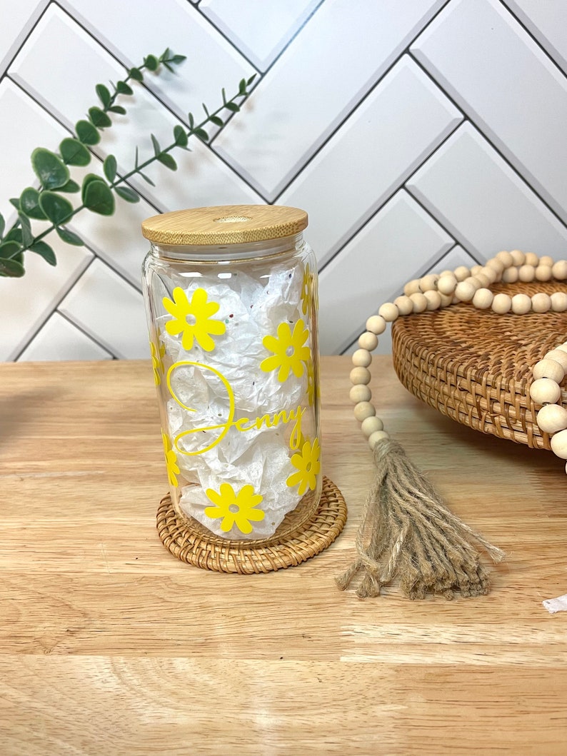 May include: A clear glass jar with a bamboo lid and yellow daisy decals. The jar is sitting on a woven coaster and has the name "Jenny" written on it.