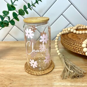 May include: A clear glass jar with a bamboo lid and pink floral design. The jar has the word "Charity" written on it. The jar is sitting on a woven coaster.