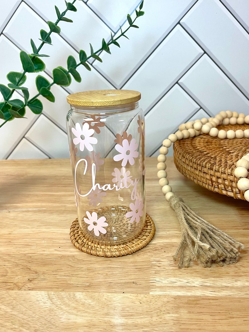 May include: A clear glass jar with a bamboo lid and pink floral design. The jar has the word "Charity" written on it. The jar is sitting on a woven coaster.