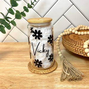 May include: A clear glass jar with a bamboo lid and black daisy decals. The jar is labeled "Vicky".