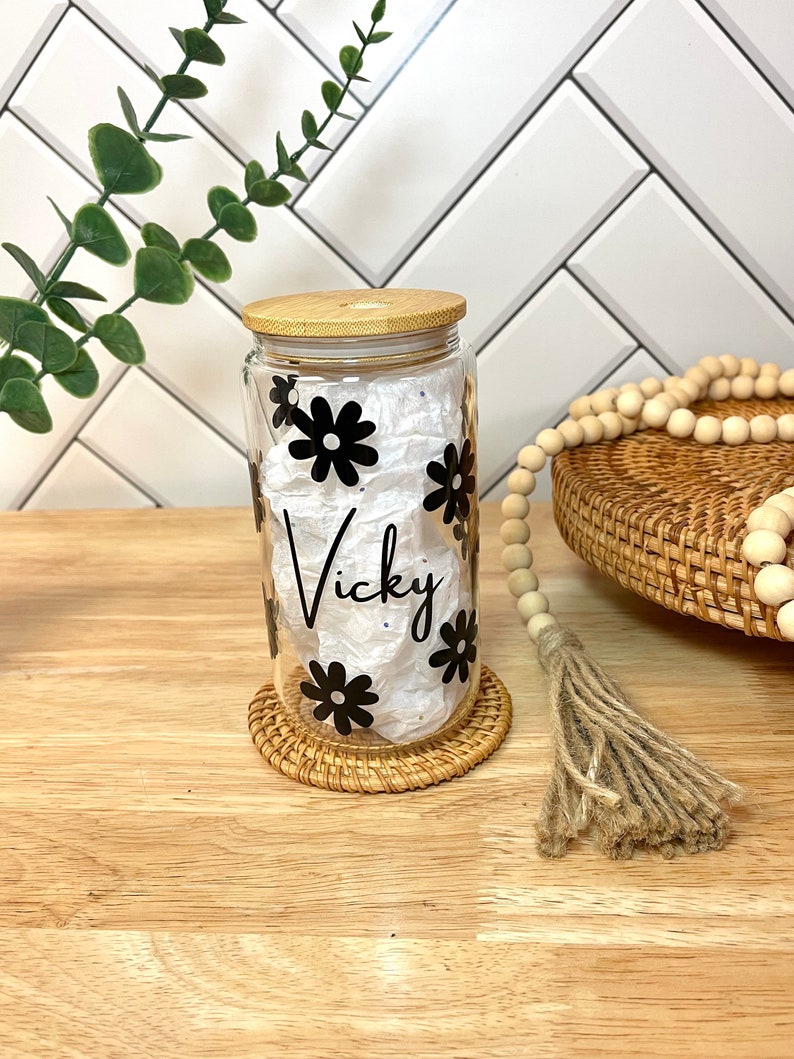 May include: A clear glass jar with a bamboo lid and black daisy decals. The jar is labeled "Vicky".