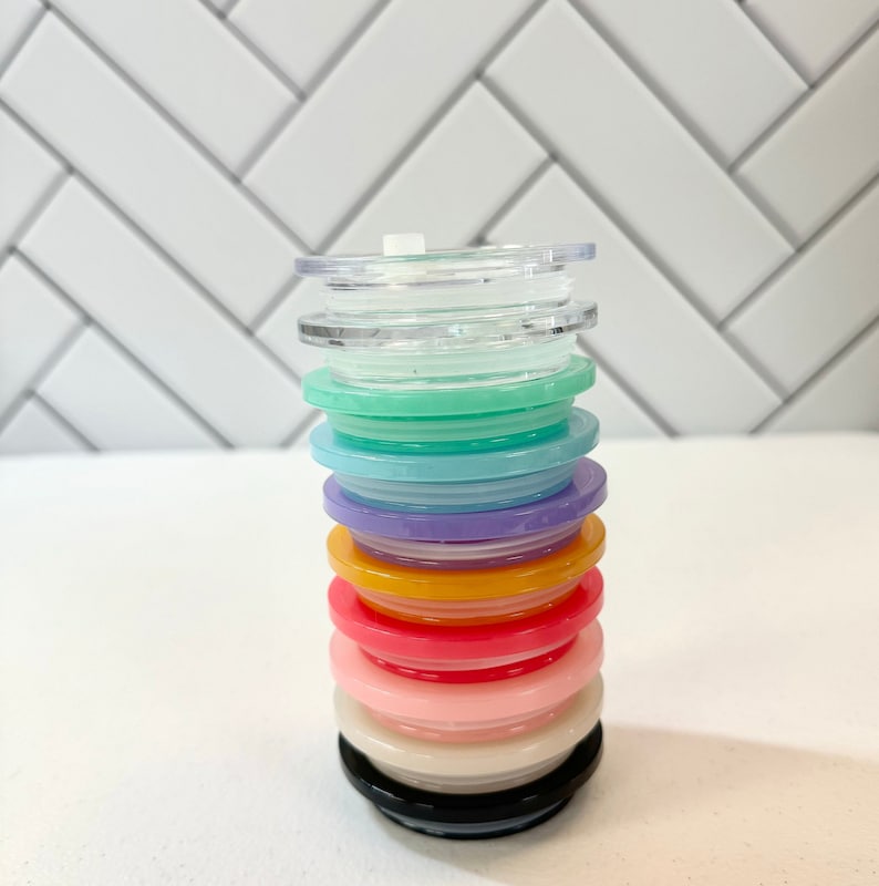 May include: A stack of colourful plastic lids for a tumbler or cup. The lids are in various colours, including pink, orange, yellow, green, blue, and purple. The lids are stacked on top of each other, with the clear lid on top.