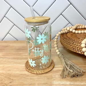 May include: A clear glass reusable water bottle with a bamboo lid and a straw. The bottle has a light blue floral design and the name "Patty" written on it. The bottle is sitting on a woven coaster.