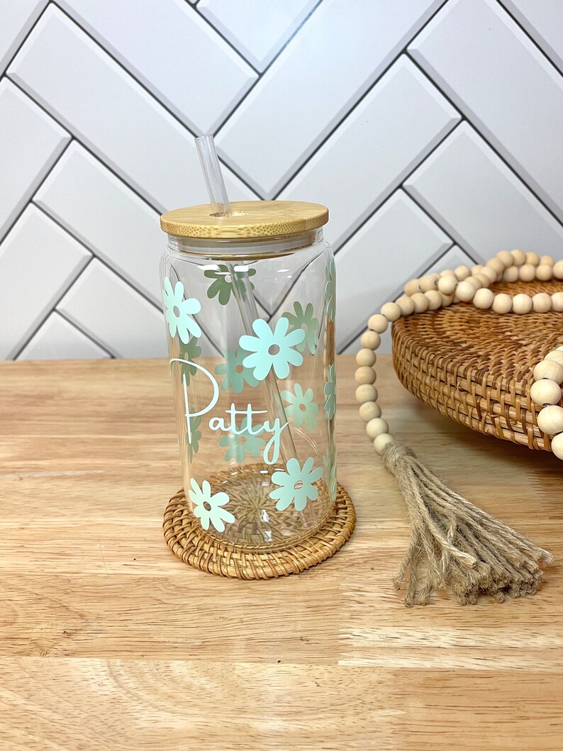 May include: A clear glass reusable water bottle with a bamboo lid and a straw. The bottle has a light blue floral design and the name "Patty" written on it. The bottle is sitting on a woven coaster.
