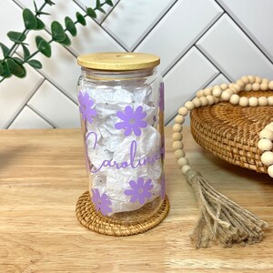 May include: A clear glass jar with a bamboo lid and purple flowers printed on the side. The jar has the name "Carolina" written on it in cursive.