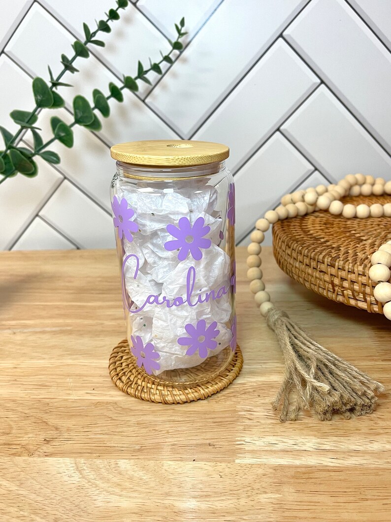 May include: A clear glass jar with a bamboo lid and purple flowers printed on the side. The jar has the name "Carolina" written on it in cursive.