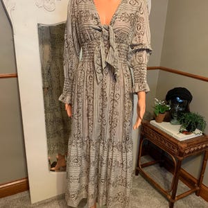 May include: A long, flowing dress with a V-neck and long sleeves. The dress features a repeating pattern in shades of brown and cream. The bodice is fitted with a tie detail at the neckline, and the skirt has a tiered design. The dress is displayed on a mannequin.