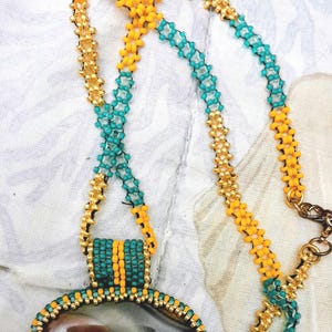May include: A necklace featuring a large, smooth, oval-shaped brown stone pendant encased in a beaded border of metallic gold and turquoise seed beads. The chain is crafted from alternating segments of yellow, turquoise, and gold seed beads.