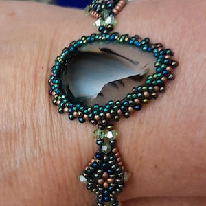 May include: A bracelet featuring a large, smooth, dark gray teardrop-shaped stone bezel-set in iridescent blue, green, and copper-colored seed beads. The band is constructed from intricate beadwork with small, light green faceted crystal accents.